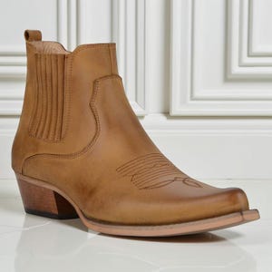 Puede incluir: Bota de vaquero de cuero marrón claro con puntera cuadrada y tacón bajo de madera. La bota presenta costuras decorativas en la pala y una lengüeta en el lateral. La bota mide unos 20 cm de alto.