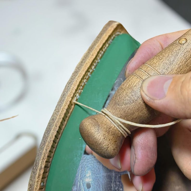 May include: Close-up of leather craft in progress. A wooden tool is used to stitch a light-colored thread through a leather edge. The leather is brown with a green interior.