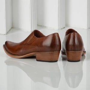 May include: A pair of brown leather cowboy boots with a pointed toe and low heel. The boots have a slip-on design with elastic side panels. The leather has a glossy finish and a decorative design on the toe.