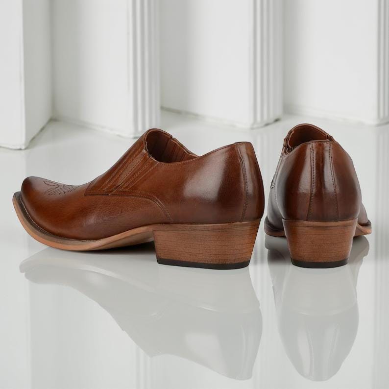 May include: A pair of brown leather cowboy boots with a pointed toe and low heel. The boots have a slip-on design with elastic side panels. The leather has a glossy finish and a decorative design on the toe.