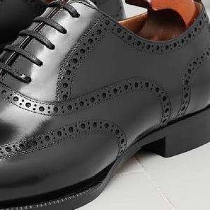 May include: Close-up of a black leather Oxford shoe with decorative perforations and a lace-up closure. The shoe has a polished finish and a stacked heel. The shoe is on a white surface.