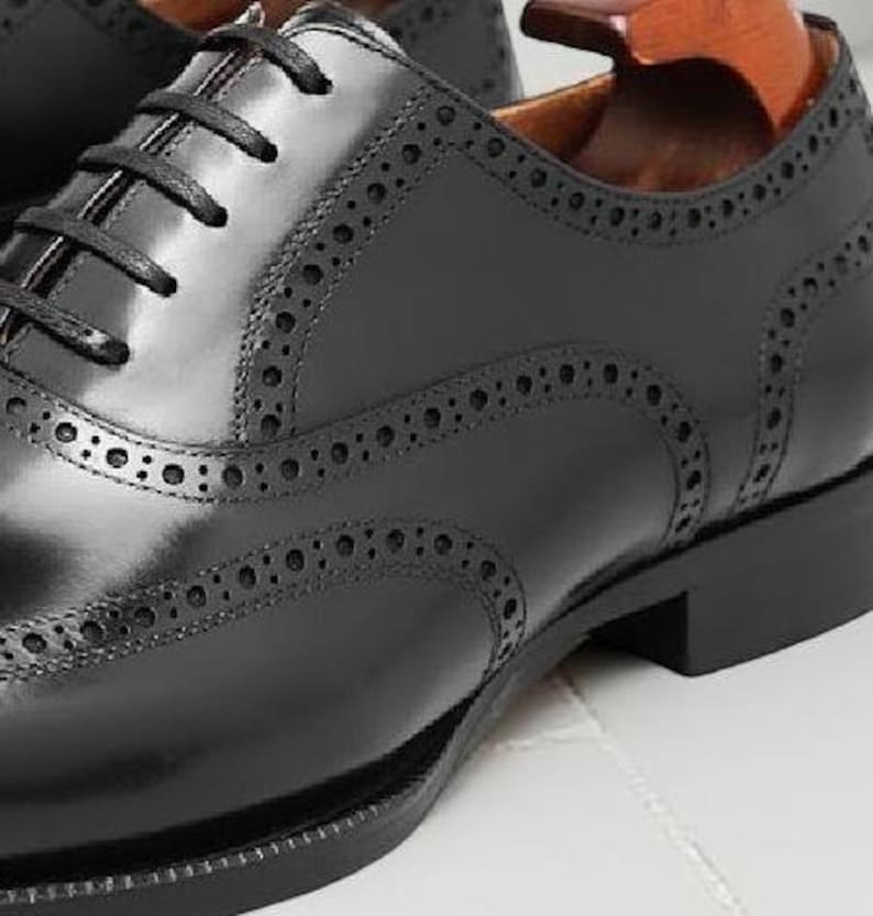May include: Close-up of a black leather Oxford shoe with decorative perforations and a lace-up closure. The shoe has a polished finish and a stacked heel. The shoe is on a white surface.
