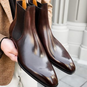 May include: A pair of polished brown leather Chelsea boots. The boots have a sleek, pointed toe design and elastic side panels. The interior lining is a light tan colour. The boots are held up by a person.