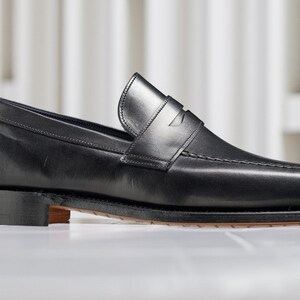 May include: A single black leather loafer shoe with a low heel. The shoe has a classic design with a strap across the top and visible stitching. The sole is a dark brown color.
