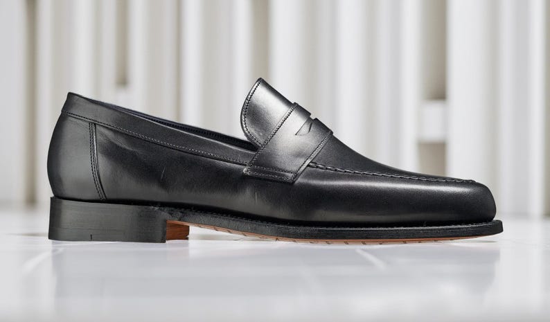 May include: A single black leather loafer shoe with a low heel. The shoe has a classic design with a strap across the top and visible stitching. The sole is a dark brown color.