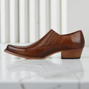 May include: A brown leather shoe with a square toe and a short, stacked heel. The shoe has a slip-on design with a stitched vamp and a subtle decorative pattern on the toe. The heel measures approximately 1.5 inches.