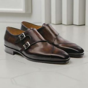 Pode incluir: Um par de sapatos castanhos de couro com fivela dupla. Os sapatos têm um acabamento polido, um design de biqueira e fivelas prateadas. Os sapatos são exibidos numa superfície branca.