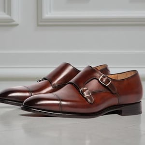 May include: A pair of brown leather double monk strap shoes. The shoes have a polished finish, a cap toe design, and silver buckles. The shoes are displayed on a light-coloured surface.