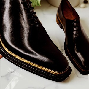 May include: A pair of black leather dress shoes with a square toe and a gold-coloured trim around the sole. The shoes have a lace-up design and are displayed on a marble surface.