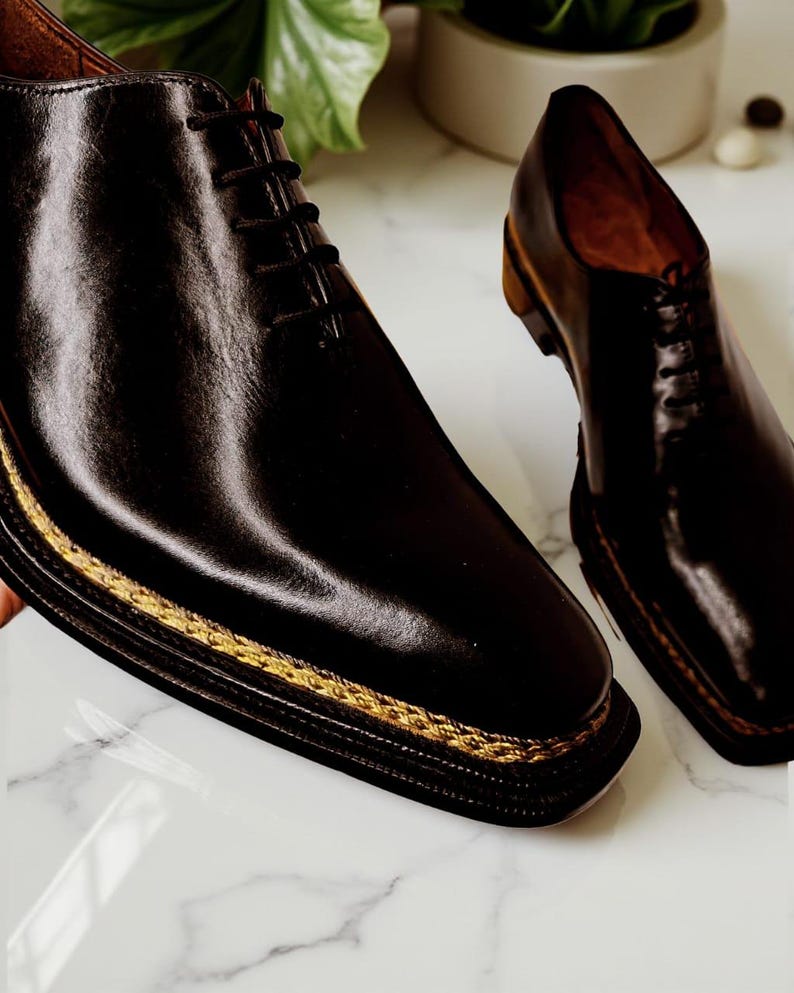 May include: A pair of black leather dress shoes with a square toe and a gold-coloured trim around the sole. The shoes have a lace-up design and are displayed on a marble surface.