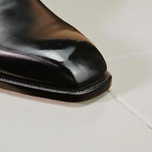 May include: Close-up of a black leather shoe with a square toe. The shoe has a glossy finish and a stitched sole. The image highlights the shoe's sleek design and craftsmanship, suitable for formal wear.