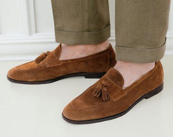 Puede incluir: Mocasines de ante marrón con borlas. Los zapatos tienen una puntera redondeada y suela marrón oscuro. Las borlas son de un tono marrón más oscuro. Pantalones verde oliva.