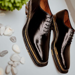 May include: A pair of black leather dress shoes with a shiny finish. The shoes feature a traditional lace-up design and a gold-coloured trim around the sole. The inside of the shoes is a light brown colour.
