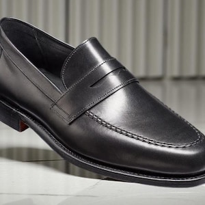 May include: A single black leather loafer shoe with a penny strap. The shoe has a rounded toe, a stitched sole, and a small heel. The shoe is displayed on a white and gray tiled surface.