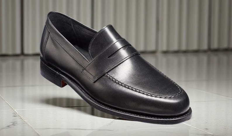 May include: A single black leather loafer shoe with a penny strap. The shoe has a rounded toe, a stitched sole, and a small heel. The shoe is displayed on a white and gray tiled surface.