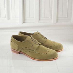 Può includere: Un paio di scarpe brogue in camoscio verde oliva con lacci marroni. Le scarpe presentano perforazioni decorative e una suola marrone chiaro. Sfondo bianco e pavimento piastrellato.