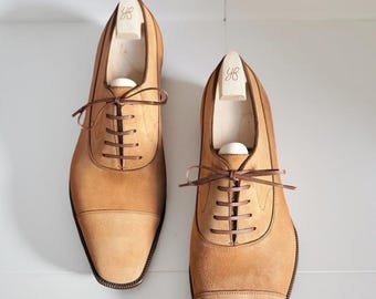 May include: A pair of tan suede dress shoes with brown laces and a cap toe design. The shoes are displayed with wooden shoe trees inside. The shoes are a light brown color and are on a white surface.