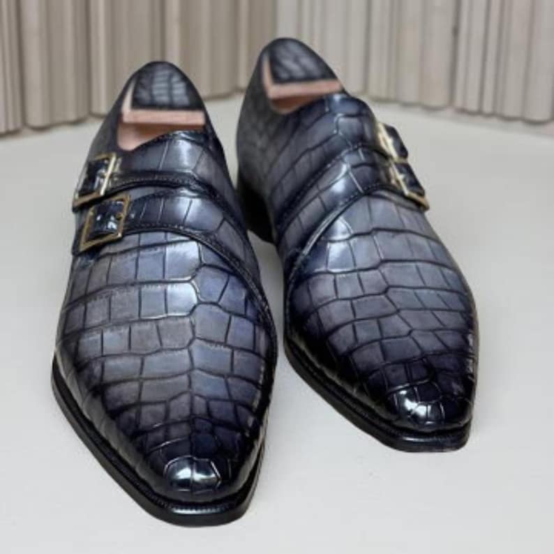 May include: A pair of gray and black leather dress shoes with a crocodile texture. The shoes have a pointed toe, double buckle closure, and a glossy finish. The buckles are gold-colored.