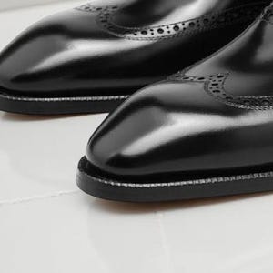 May include: Close-up of a pair of black leather dress shoes. The shoes have a glossy finish, decorative perforations, and a stitched sole. The image highlights the craftsmanship and detail of the footwear.