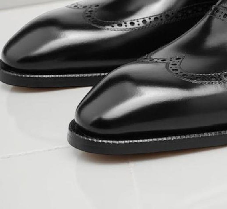 May include: Close-up of a pair of black leather dress shoes. The shoes have a glossy finish, decorative perforations, and a stitched sole. The image highlights the craftsmanship and detail of the footwear.