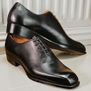 May include: A pair of black leather dress shoes with a square toe and lace-up closure. The shoes have a polished finish and a stacked heel, suitable for formal occasions.