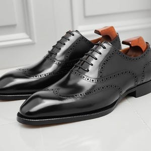 May include: A pair of black leather Oxford shoes with brogue detailing. The shoes have a glossy finish, a lace-up closure, and wooden shoe trees inside. The shoes are displayed on a white surface.
