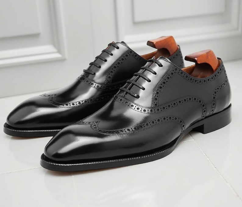 May include: A pair of black leather Oxford shoes with brogue detailing. The shoes have a glossy finish, a lace-up closure, and wooden shoe trees inside. The shoes are displayed on a white surface.