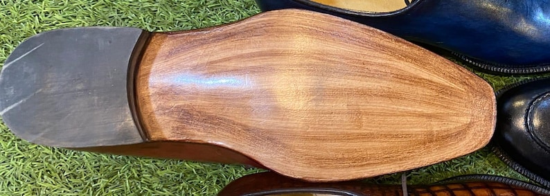 May include: Close-up of the sole of a brown leather shoe with a dark gray heel. The shoe's sole has a wood-grain texture. Other leather shoes in blue and black are visible in the background.