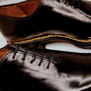 May include: A pair of black leather dress shoes with a glossy finish. The shoes have black laces and a gold-coloured trim along the sole. The interior of the shoe is a light brown colour.