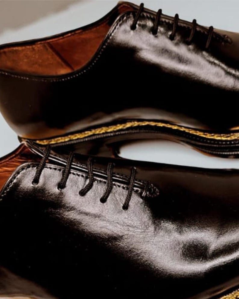 May include: A pair of black leather dress shoes with a glossy finish. The shoes have black laces and a gold-coloured trim along the sole. The interior of the shoe is a light brown colour.