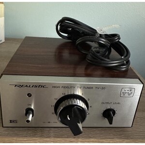 Puede incluir: Un sintonizador de TV de alta fidelidad Realistic TV-20 vintage con una parte superior de vetas de madera y una cara plateada. Incluye un dial de sintonización, control de nivel de salida e indicador de TV VHF. Un cable de alimentación negro está enrollado encima.