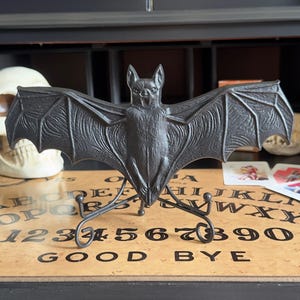 May include: A black metal bat sculpture with outstretched wings, mounted on a decorative stand. The bat is detailed with textured wings and a menacing expression. The sculpture is displayed on a wooden Ouija board.