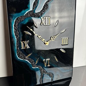 Può includere: Orologio da parete rettangolare con un design nero, verde acqua e argento. Il quadrante dell'orologio presenta numeri romani e lancette color oro. Un design strutturato nero a forma di albero borda il lato sinistro e inferiore dell'orologio.