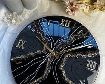 Peut inclure: Une horloge murale ronde noire avec des chiffres romains et des aiguilles dorées. Le cadran de l'horloge présente un motif doré et blanc avec une finition brillante. L'horloge est entourée de bougies et de cristaux décoratifs.