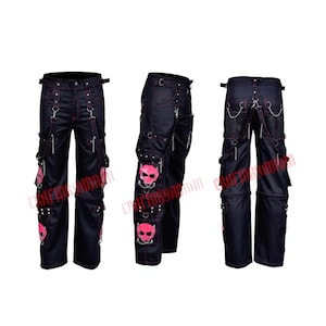 Puede incluir: Pantalones vaqueros negros con costuras y detalles en rosa. Los pantalones tienen múltiples bolsillos, cadenas de metal y parches de calaveras rosas. El diseño incluye trabillas para tirantes y herrajes metálicos decorativos.