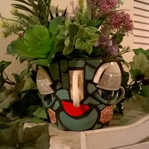 May include: A mosaic planter shaped like a face with a red mouth, blue eyes, and a white nose. The planter is filled with green plants and flowers.
