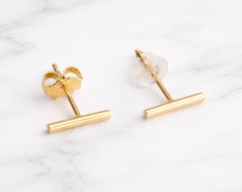 May include: A pair of gold-toned bar stud earrings. Each earring features a horizontal metal bar attached to a post. One earring is shown from the front, while the other is shown from the side with a clear plastic earring back attached to the post.