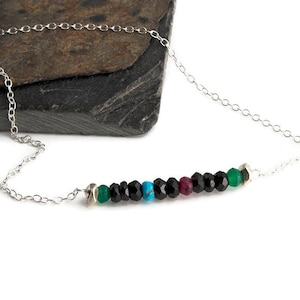 May include: A delicate silver necklace featuring a row of colorful beads. The beads include green, black, turquoise, and red gemstones. The necklace is displayed on a dark, textured surface.