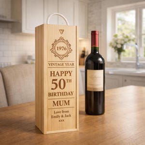 Peut inclure: Un coffret à vin en bois clair avec une poignée en corde et le texte gravé "1976 Année Vintage Joyeux 50e Anniversaire Maman". Une bouteille de vin rouge avec un bouchon rouge est à côté du coffret.