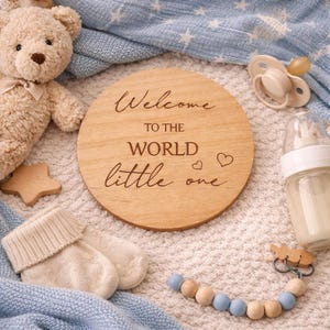 May include: A wooden disc with the text "Welcome to the world little one" is surrounded by baby items. A teddy bear, pacifier, bottle, baby socks, and a beaded pacifier clip are visible. The items are on a white and blue blanket.