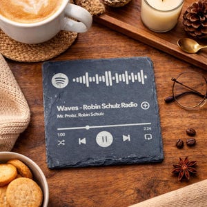 Può includere: Un sottobicchiere in ardesia nera con un codice Spotify e il testo "Waves - Robin Schulz Radio". Il sottobicchiere mostra le informazioni sulla canzone e i controlli di riproduzione. È su una superficie di legno, insieme a una tazza di caffè e altri oggetti.
