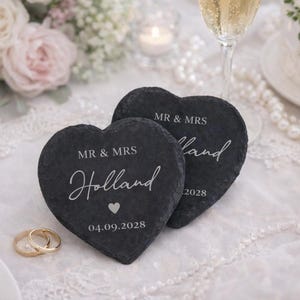 Può includere: Due sottobicchieri a forma di cuore in ardesia con la scritta "MR & MRS Holland" e la data "04.09.2028". I sottobicchieri sono neri con scritte bianche e un piccolo cuore. Tema matrimonio con fedi d'oro e fiori.