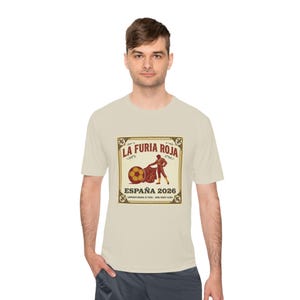 May include: A light beige t-shirt featuring a graphic with the text "LA FURIA ROJA ESPAÑA 2026". The graphic depicts a matador with a red cape and a soccer ball. The shirt is made of a soft, comfortable material.