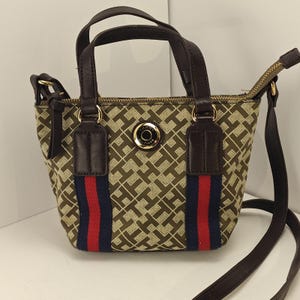 May include: A small, brown handbag with a repeating pattern of interlocking "H" shapes. The bag features dark brown handles and a detachable shoulder strap. A red and blue stripe runs down the front. The bag has a gold zipper and a gold emblem.