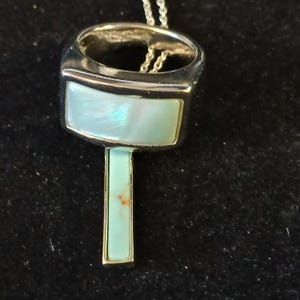 May include: A silver-toned pendant necklace with a key-shaped design. The pendant features a rectangular, iridescent, light blue stone set within a silver frame. The key's handle is a matching light blue stone. The necklace is on a silver chain.