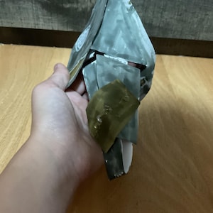 May include: A handmade paper sculpture, primarily gray and white, with a folded, angular design. A section of olive green paper is attached. The sculpture is held in a hand, with the background showing a wooden floor and a dark wooden surface.