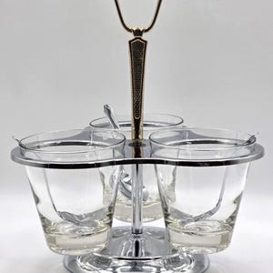 May include: A vintage serving set with three clear glass bowls, each with a small spoon. The bowls are held in a chrome-colored metal frame with a gold-colored handle. The base is also chrome-colored.