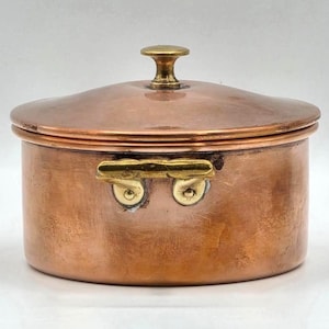 May include: A round copper pot with a matching lid and a brass knob handle. The pot features two brass side handles attached with rivets. The exterior surface of the copper shows a polished finish with natural variations in the metal color.