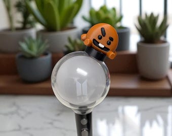 May include: A black and silver light stick with a clear, spherical head featuring the BTS logo. An orange cartoon character is perched on top. The background includes several potted succulents.