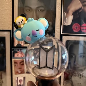 May include: A clear, spherical light stick with a black base, featuring a white and black logo. Adorned with a light blue koala and yellow character. The background includes framed portraits. The light stick is held in a hand.